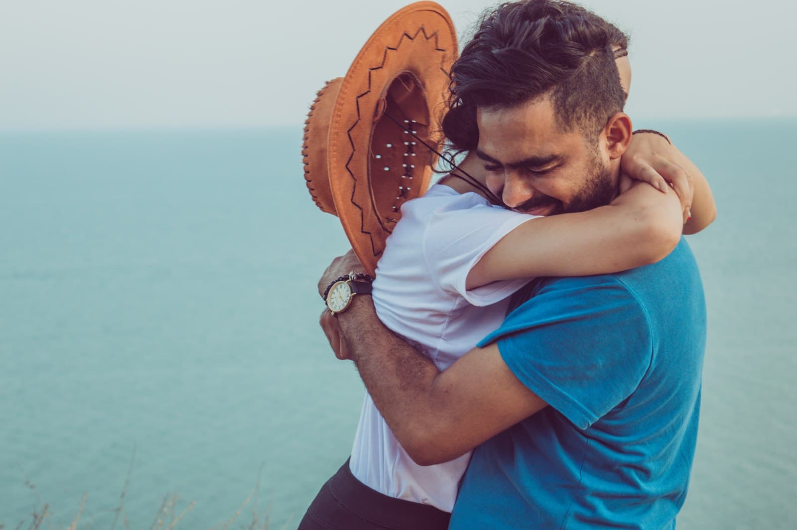 A young couple hugging on a clifftop above a calm turquoise sea, the man smiling